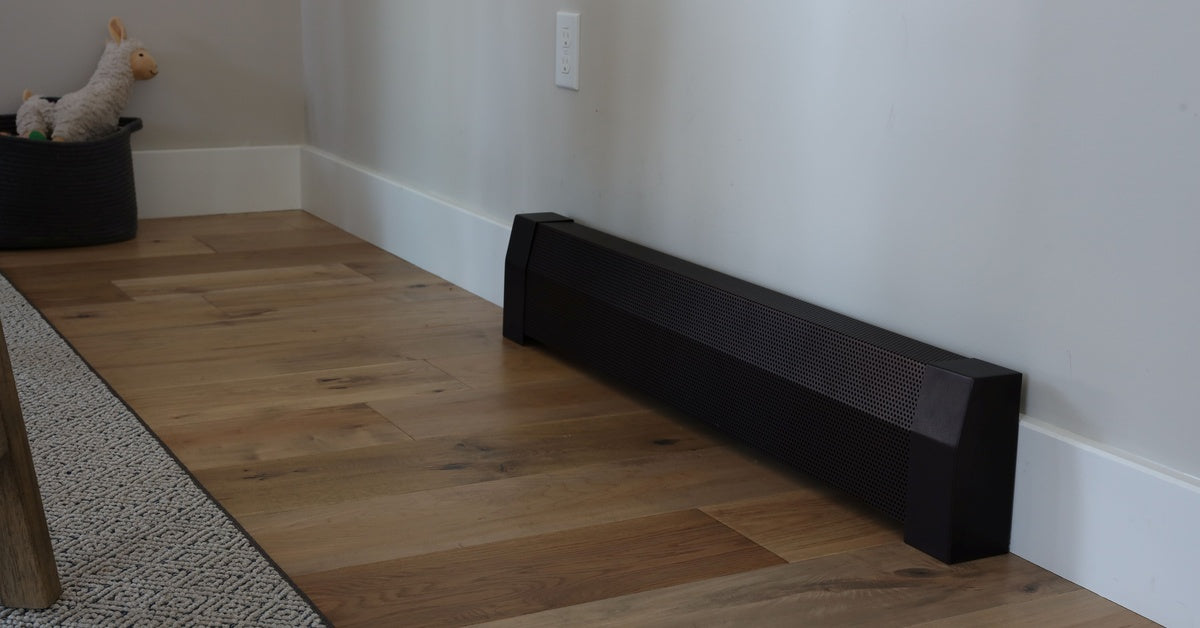 A long, dark baseboard heater sitting against a white wall on a wooden floor beside a gray rug and toy basket.