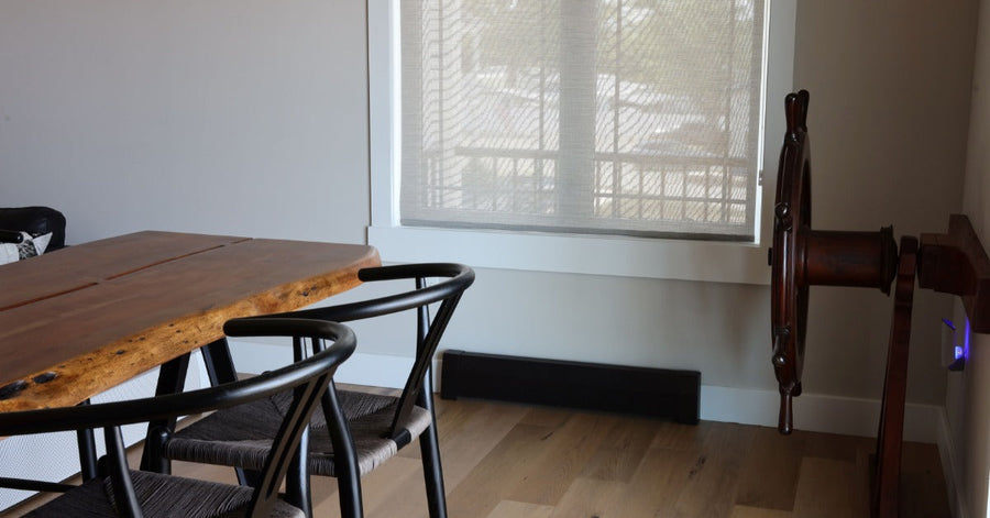 A modern dining area with a wooden table, black chairs, large window, baseboard heater, and a wooden ship wheel on the wall.