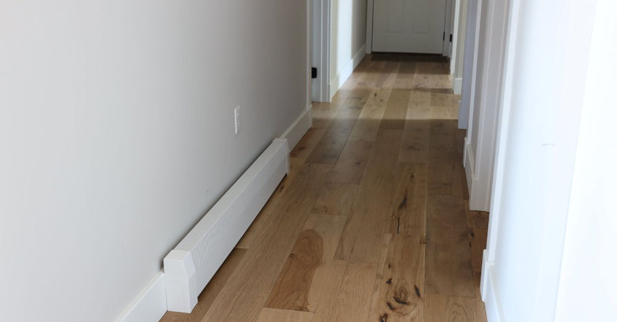 A bright hallway with hardwood flooring, white trim, multiple doorways, and a white baseboard heater cover.