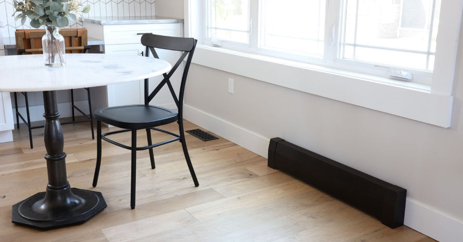 A bright dining area with a round table, black chair, large windows, and a black baseboard heater cover.